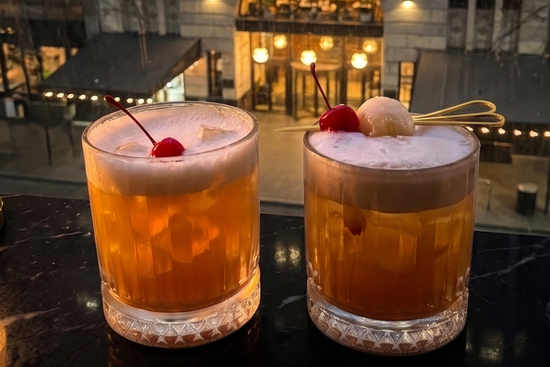 Two amber cocktails with white foam and cherry garnishes sit on a dark bar counter in front of a softly lit city building at dusk.